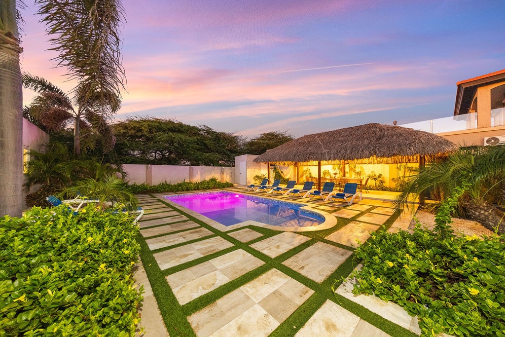 An outdoor courtyard features a pristine swimming pool with colorful lighting, surrounded by tiled pathways and lush greenery. A thatched-roof gazebo offers shaded seating, while sun lounges provide space for relaxation under the evening sky.