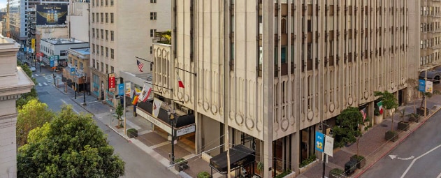 The exterior of a modern building showcases distinct architectural lines and patterns. Flags wave gently, contributing to the vibrant atmosphere of the street below. Green trees line the sidewalk, adding a touch of nature to the urban setting.