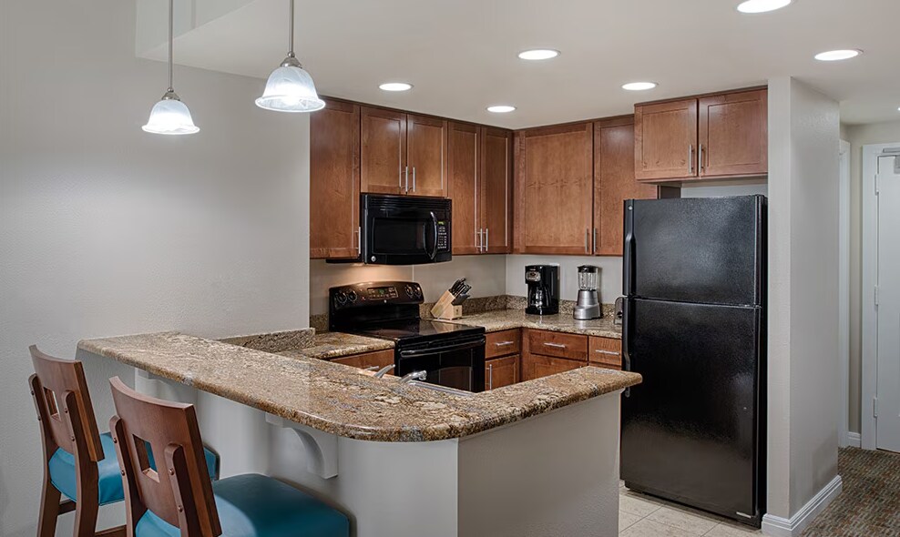 A modern kitchen features wooden cabinetry and a granite countertop. Black appliances, including a refrigerator and stove, are visible. Two bar stools with teal cushions are positioned at the counter, and recessed lighting illuminates the space.