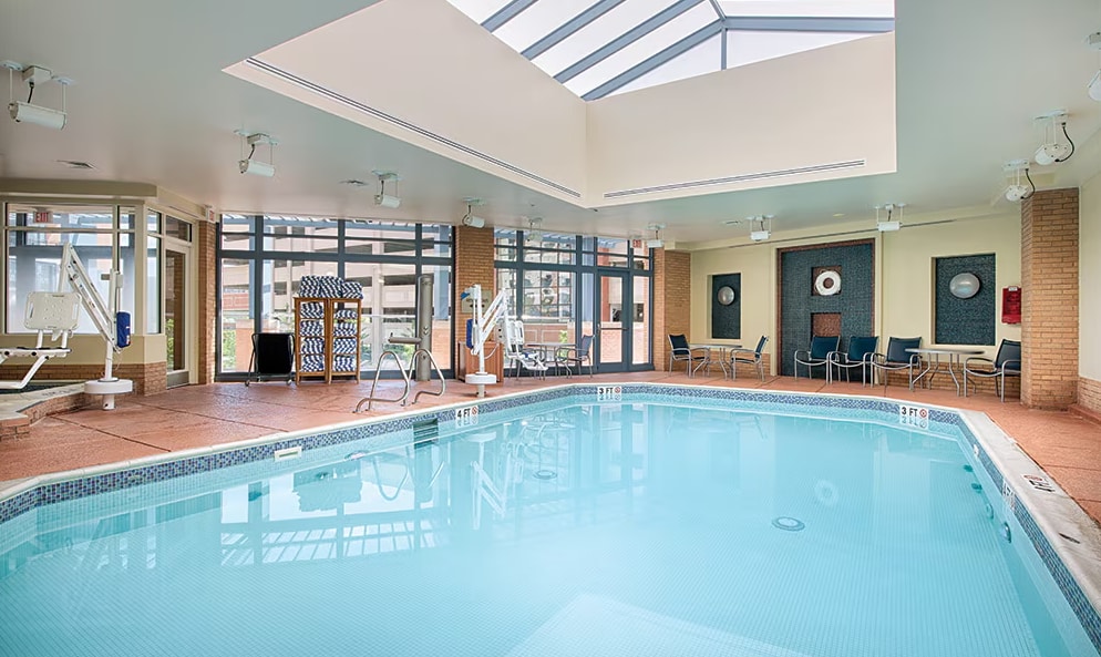 An indoor swimming pool is illuminated by natural light filtering through the skylights. Seating areas with chairs are arranged around the pool, and a shallow end is visible. The walls feature large windows, enhancing the airy atmosphere of the space.
