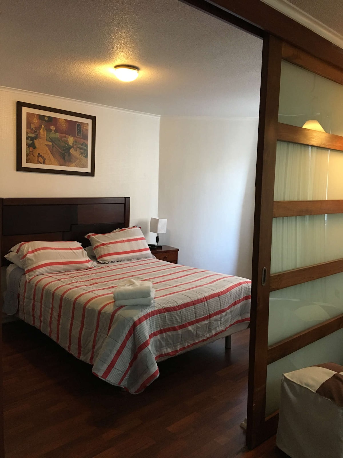 A cozy bedroom features a double bed with a striped blanket, flanked by two bedside tables with lamps. A piece of artwork adorns the wall above the bed. Natural light enters through a glass sliding door, revealing a simple and welcoming atmosphere.