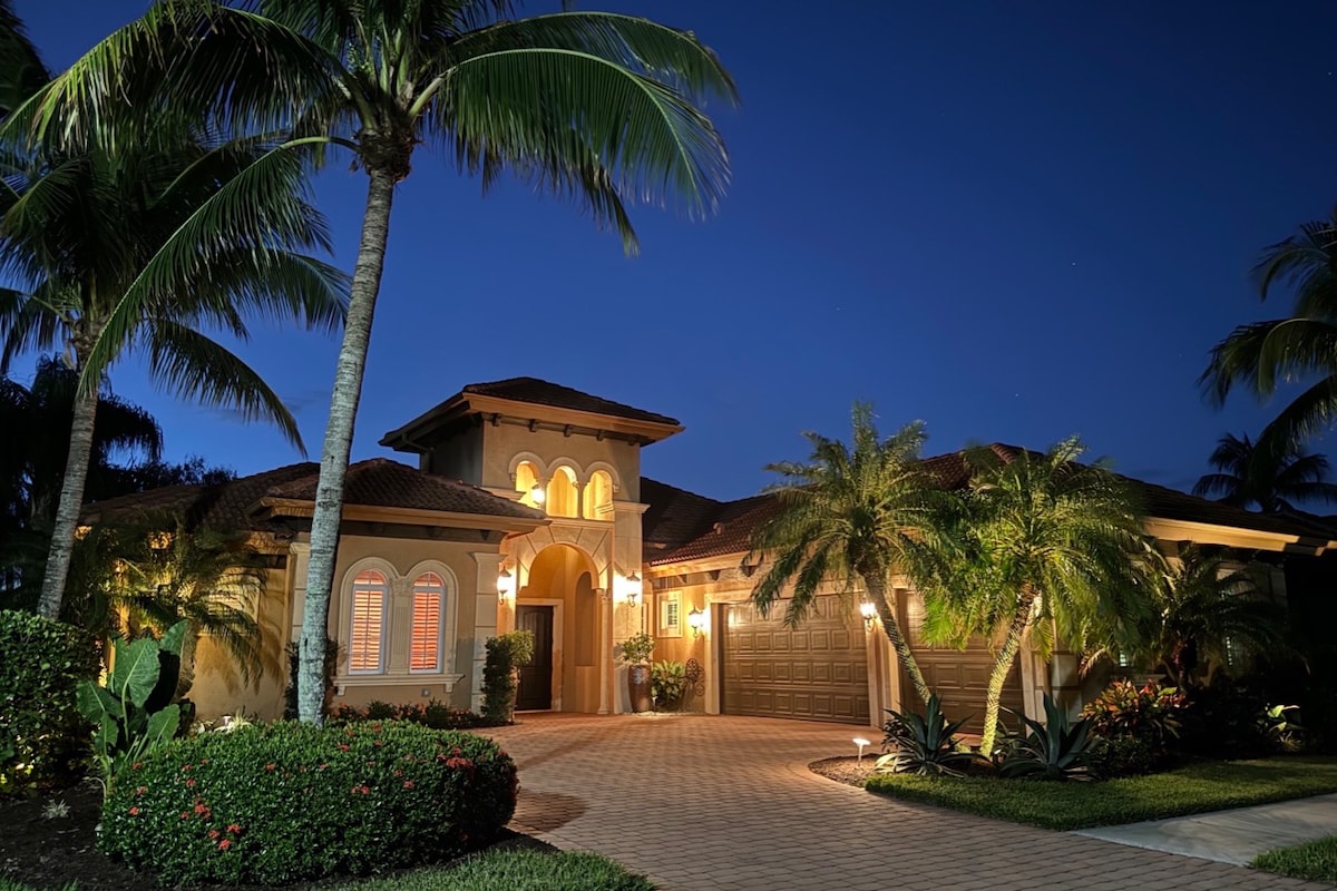 The home is viewed from the driveway at night, featuring a welcoming façade with soft lighting. Tropical palm trees frame the entrance, where elegant arches and decorative windows add charm. The driveway is paved, leading to a garage, while lush landscaping surrounds the property.