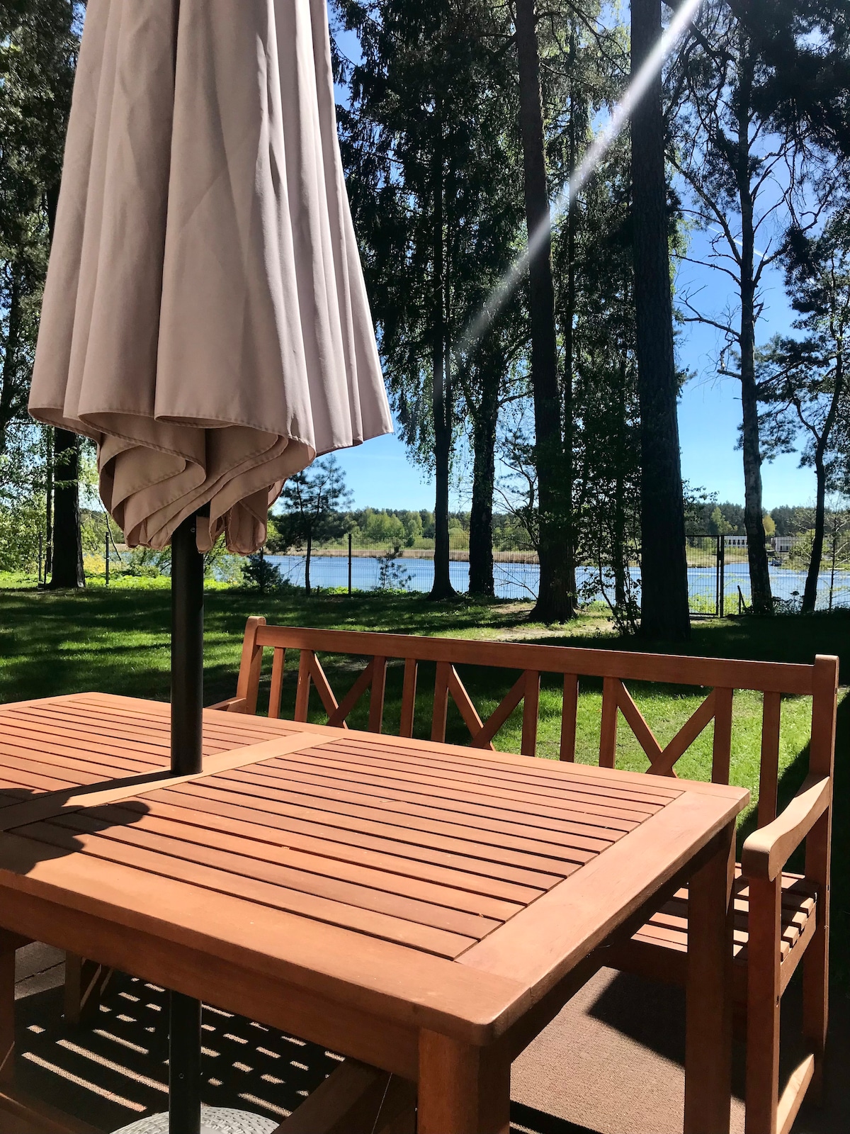 A wooden table and benches are situated on a terrace, shaded by an umbrella. Lush greenery surrounds the area, with tall trees framing a view of the river in the background under a clear blue sky.