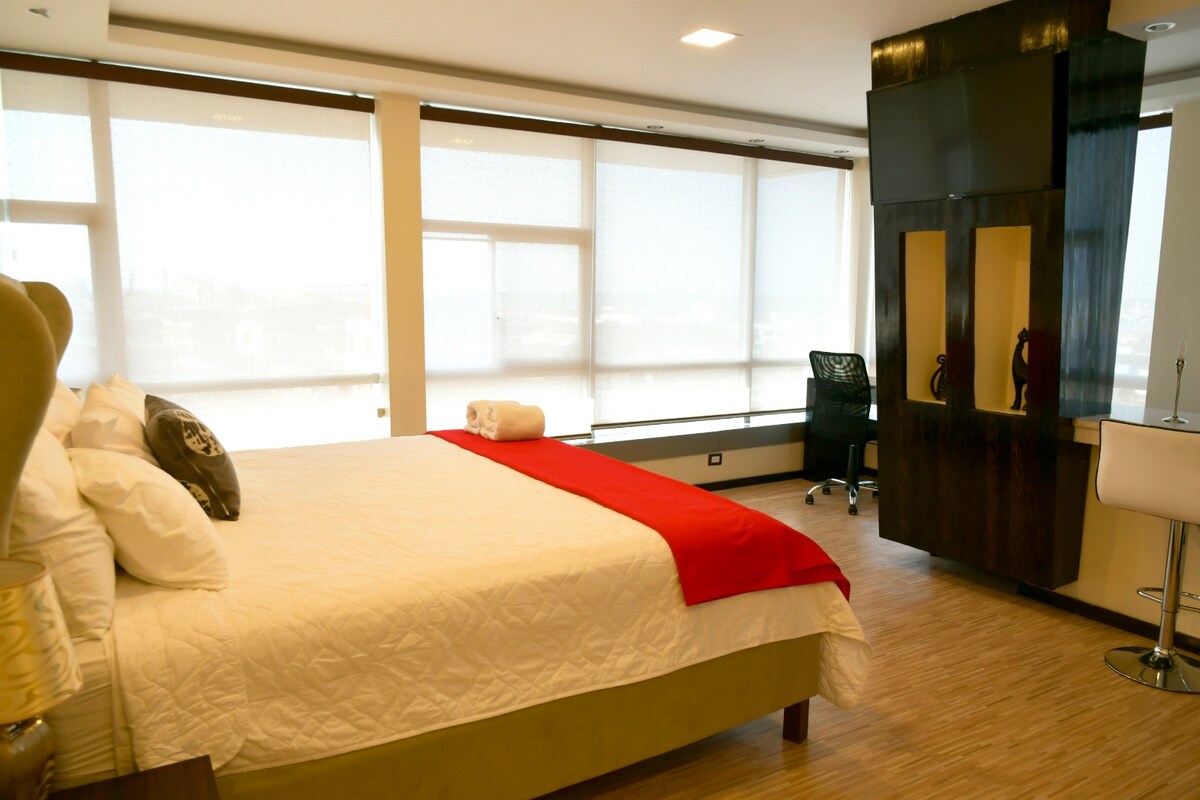 A spacious bedroom is adorned with a large bed featuring a white quilt and red throw. Natural light filters through expansive windows, revealing a modern work area with a desk and chair. A sleek cabinet with a TV is positioned adjacent to the bed.