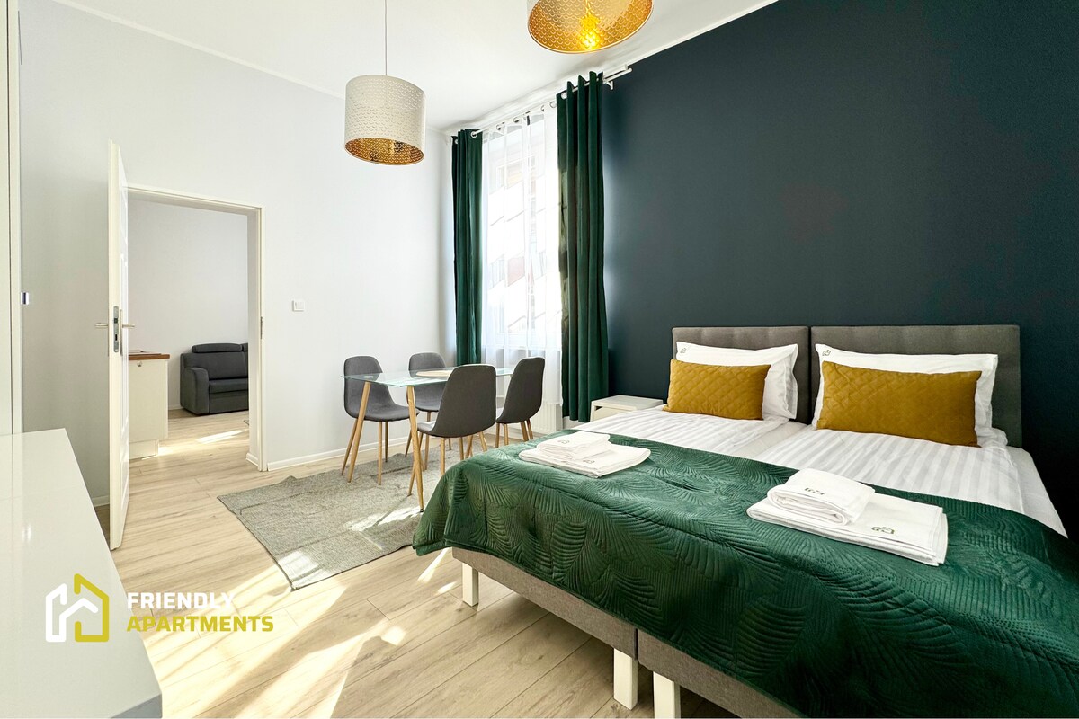 A spacious bedroom is highlighted by a dark green accent wall, featuring a comfortable bed adorned with neatly arranged white linens and yellow accent pillows. A dining area with a round table and four chairs is visible, complemented by natural light streaming through large windows with green curtains.
