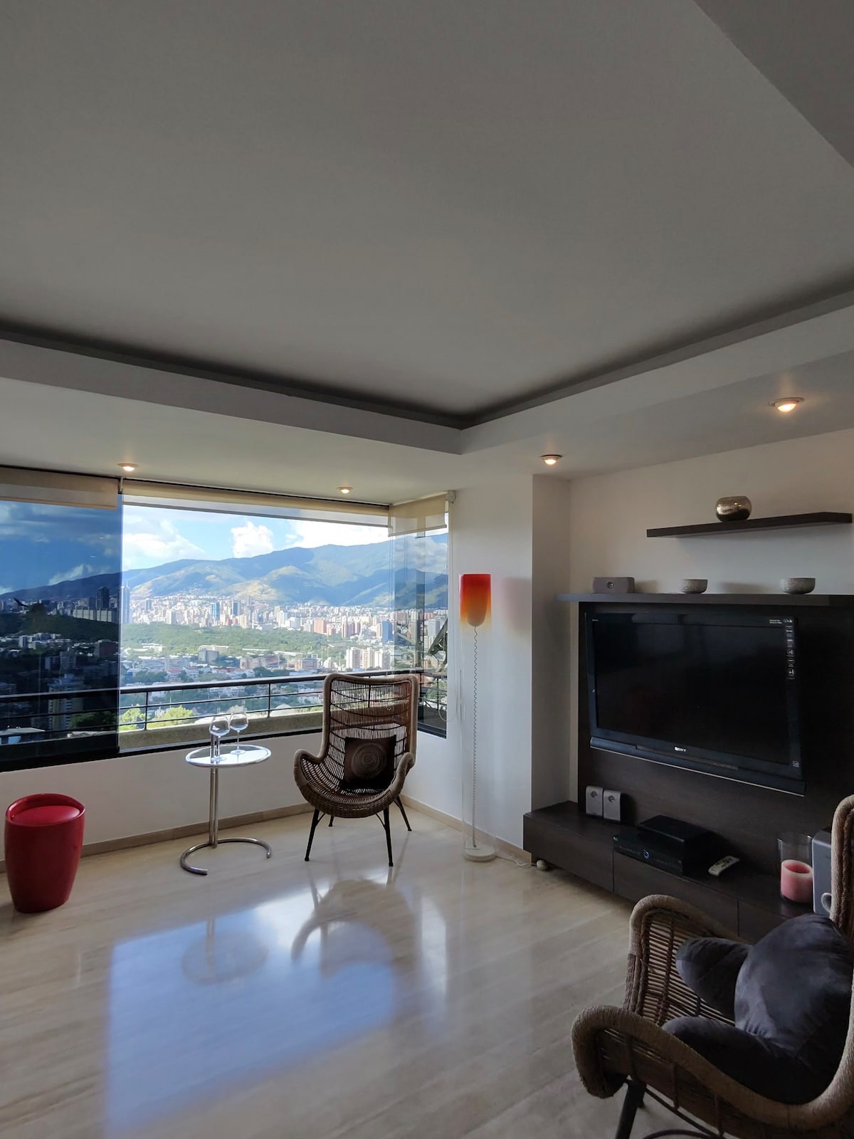 A spacious seating area features a stylish chair and a small glass table. Large windows provide panoramic views of surrounding mountains and the city below. A modern entertainment unit is visible along the wall, complemented by subtle lighting and a decorative element.