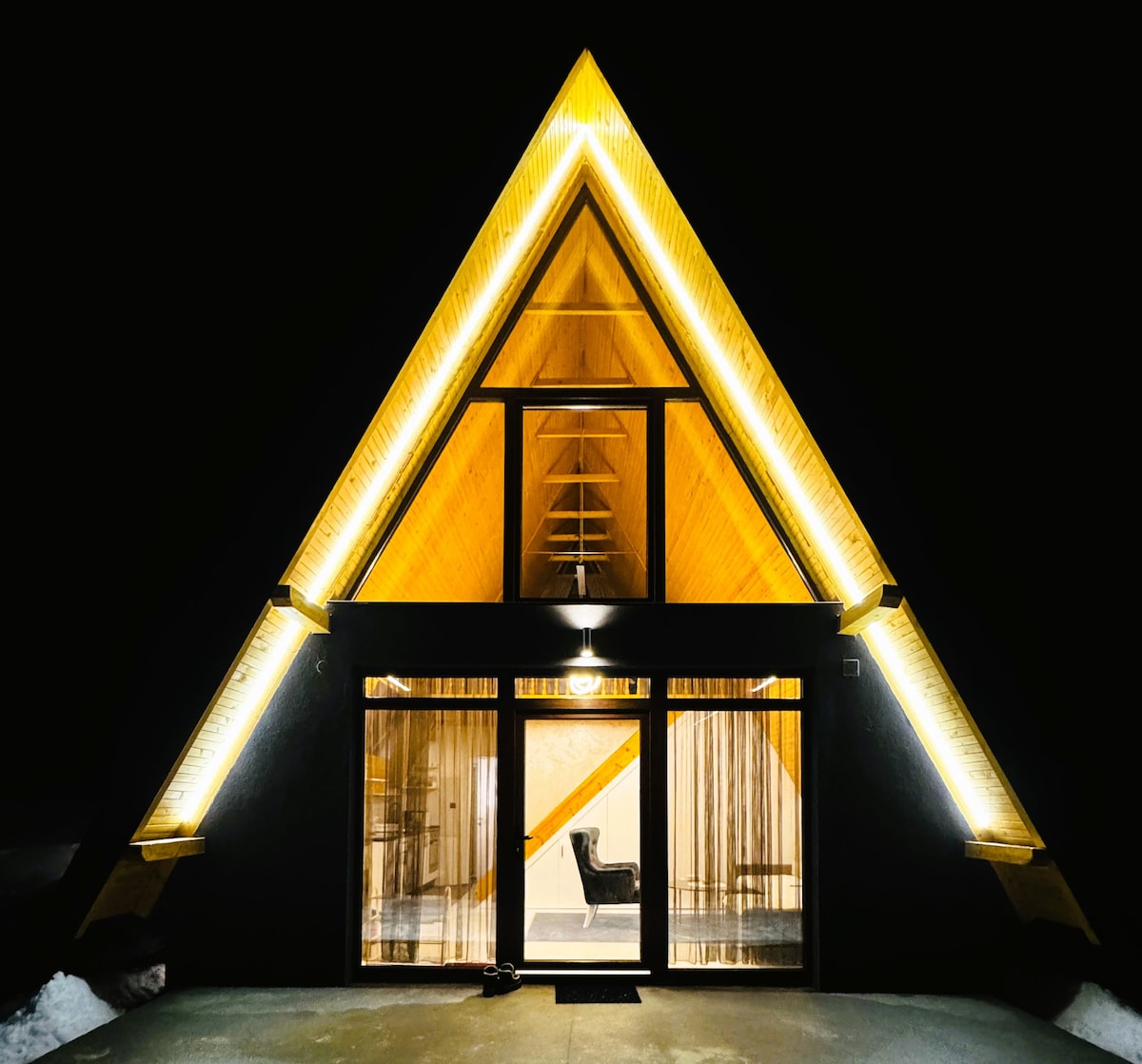 The exterior of the A-frame house is illuminated by warm lights, highlighting its striking triangular shape. Large glass doors reflect the soft glow, showcasing a welcoming entrance. The surrounding area features a clear path leading up to the front door, with snow visible on the ground.