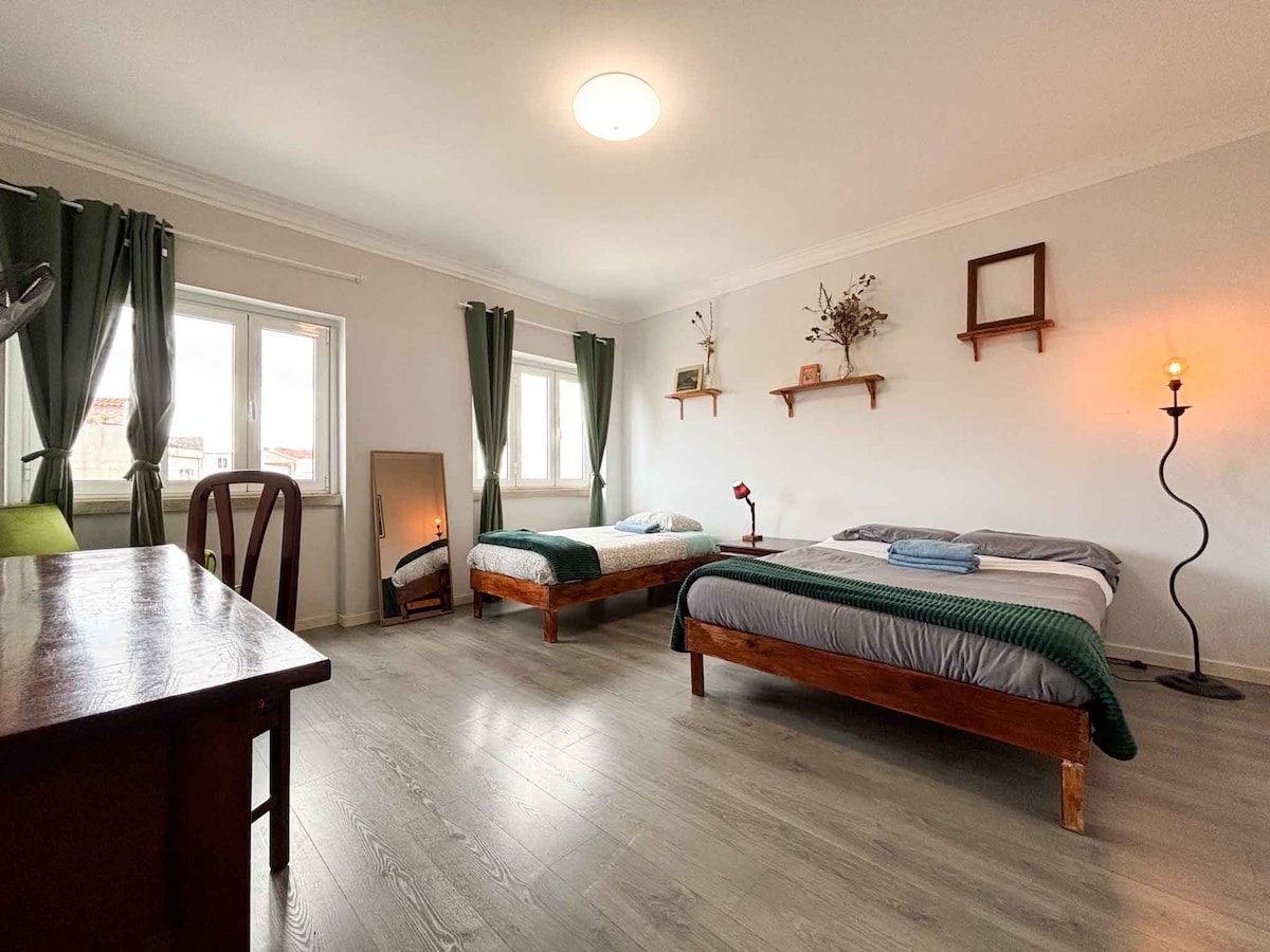 A spacious room is illuminated by soft lighting and natural light from the large windows. It features a cozy double bed and a single bed, both made up with simple bedding. A wooden desk is positioned nearby, along with decorative shelves and a floor lamp.