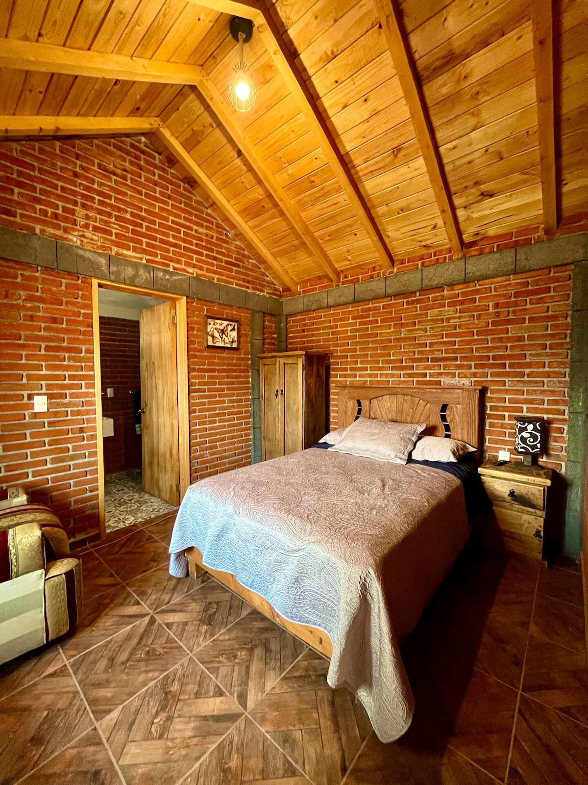 A cozy sleeping area features a spacious bed with a muted blanket and decorative pillows. Exposed wooden beams and brick walls add texture, while natural light fills the room through a nearby door. A bedside table stands beside the bed, and a comfortable chair is placed in the corner.