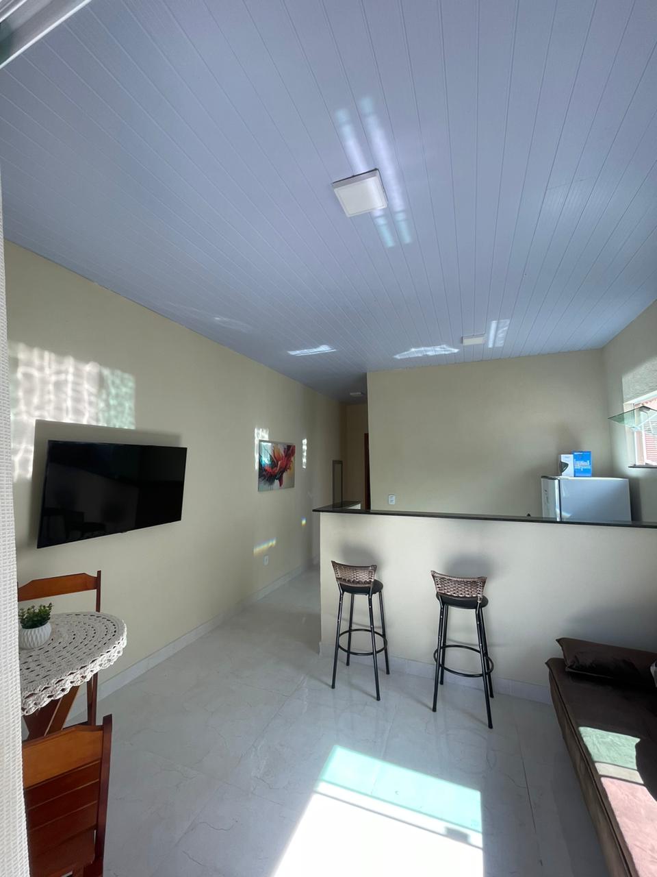 A bright living space features light-colored walls and a modern ceiling. A wall-mounted television is visible alongside a small dining area with a round table and two chairs. A kitchenette is seen in the background, with a refrigerator and minimal countertop space. Natural light filters through the room.
