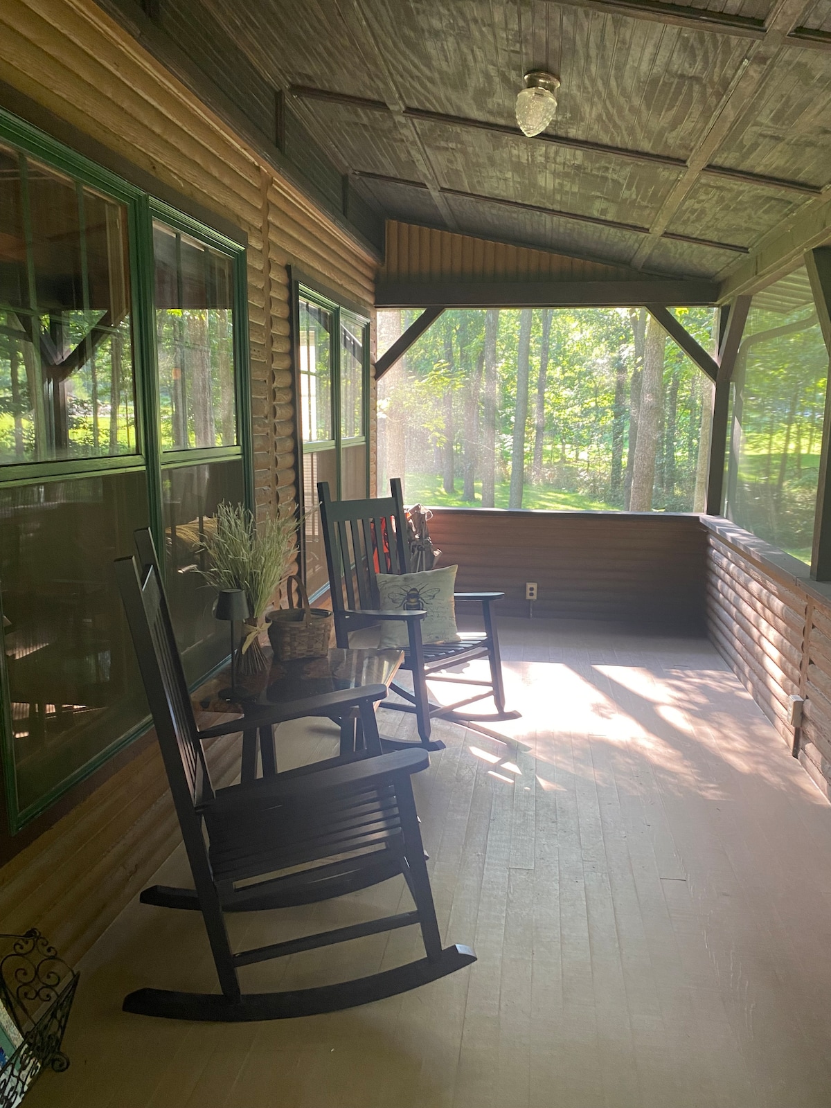 A screened porch features two black rocking chairs positioned beside a small table. Natural light filters through large windows, illuminating the wooden walls and floor. Potted plants are placed on the table, enhancing the serene atmosphere of the outdoor space.
