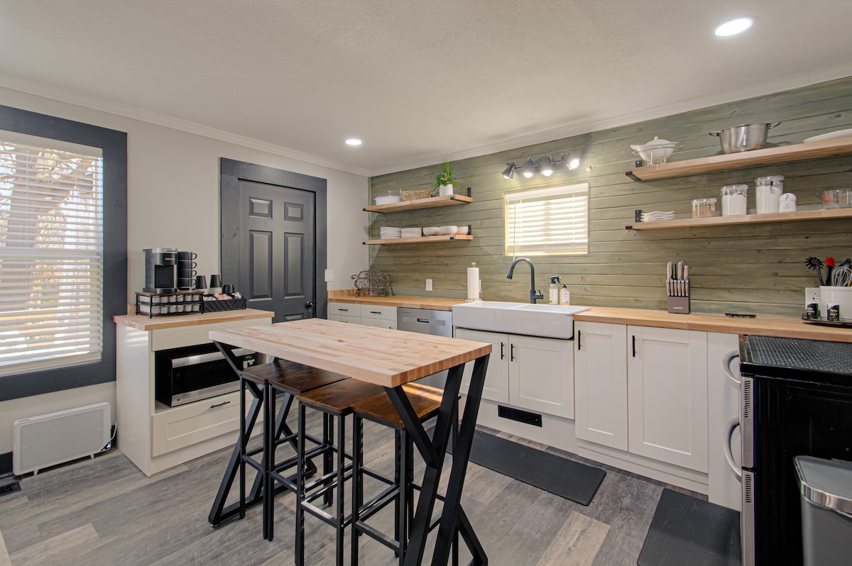 The kitchen features a minimalist design with light-colored cabinetry and wood countertops. Open shelving displays kitchenware and utensils. A central island with high stools provides additional workspace. Natural light enters through a window, enhancing the functional layout.