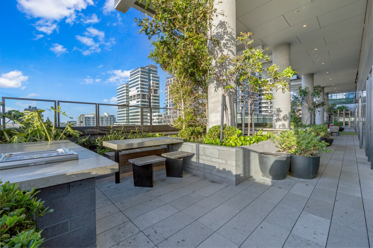 An outdoor terrace features modern landscaping with greenery and trees. A stone barbecue area is visible alongside wooden seating. The tiled surface is bordered by planters, providing a serene urban space with views of the surrounding city buildings and blue sky.