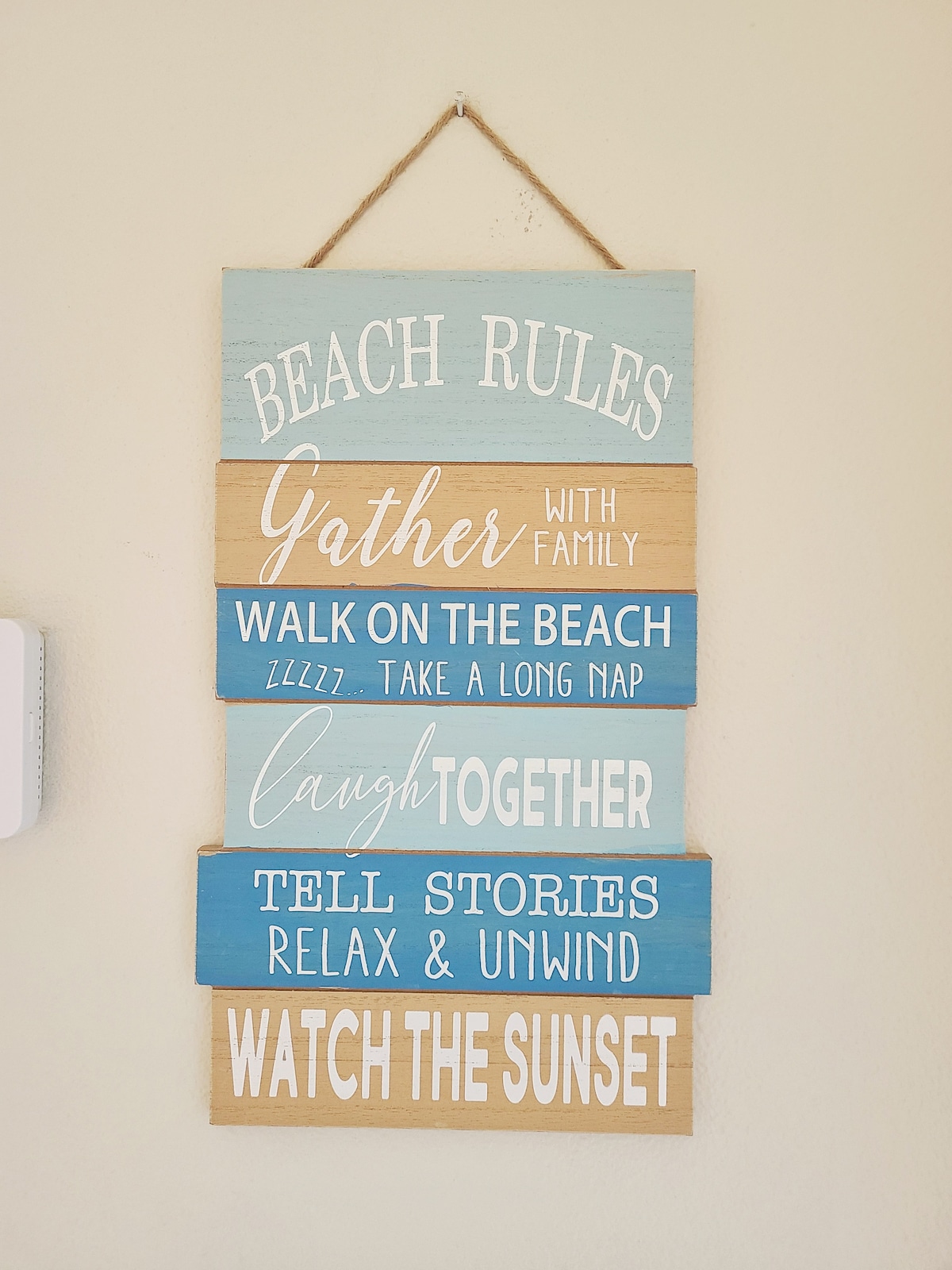 A wooden sign displaying beach rules is hung on a wall. The sign features a mix of colors, including soft blue, tan, and white, with phrases such as 'Gather with family' and 'Watch the sunset' painted in various fonts, inviting relaxation and enjoyment.