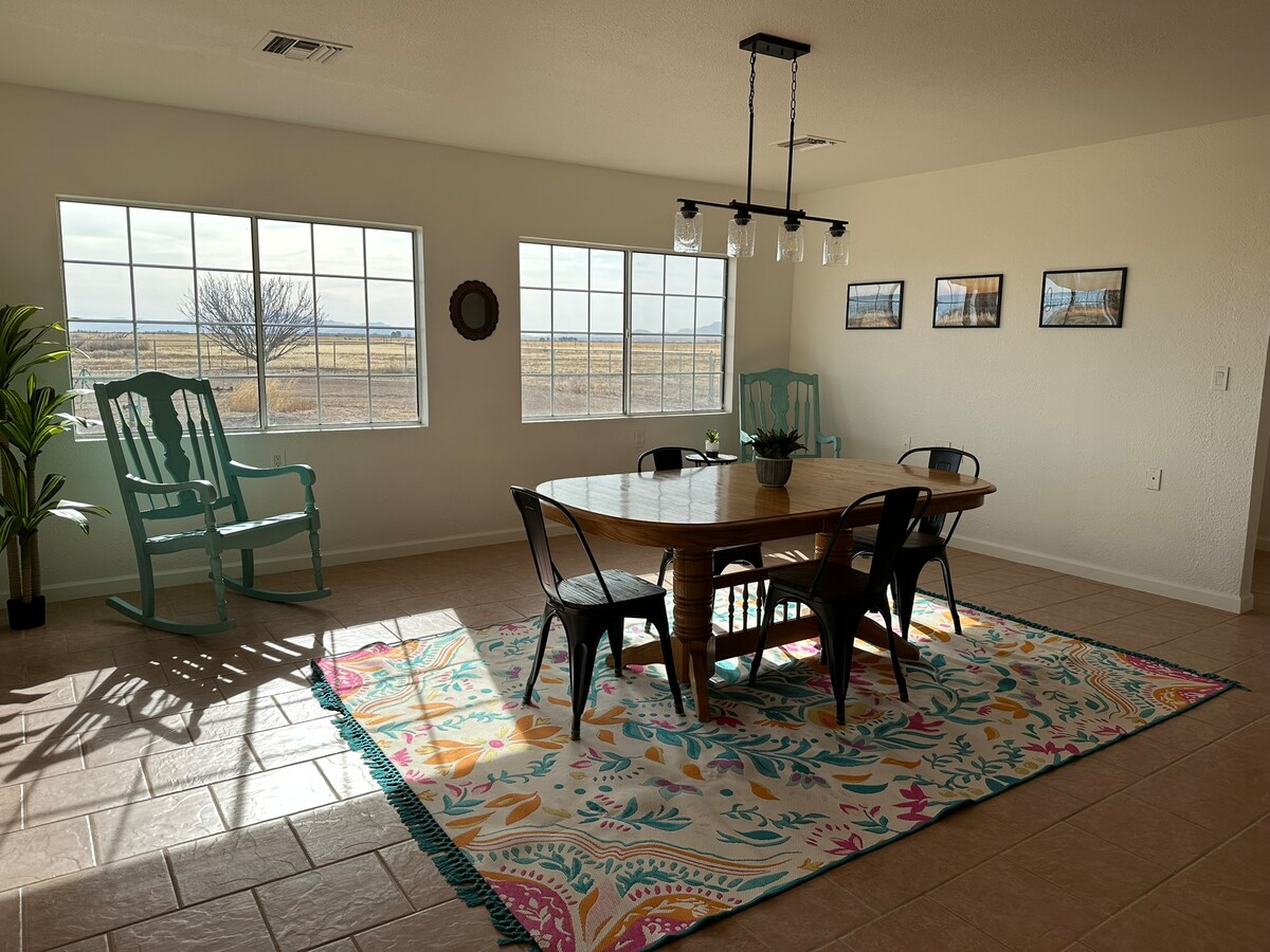 A bright dining area features a wooden table surrounded by black metal chairs. Sunlight filters through expansive windows, illuminating a colorful area rug below. Two turquoise rocking chairs are positioned nearby, enhancing the room's inviting feel.