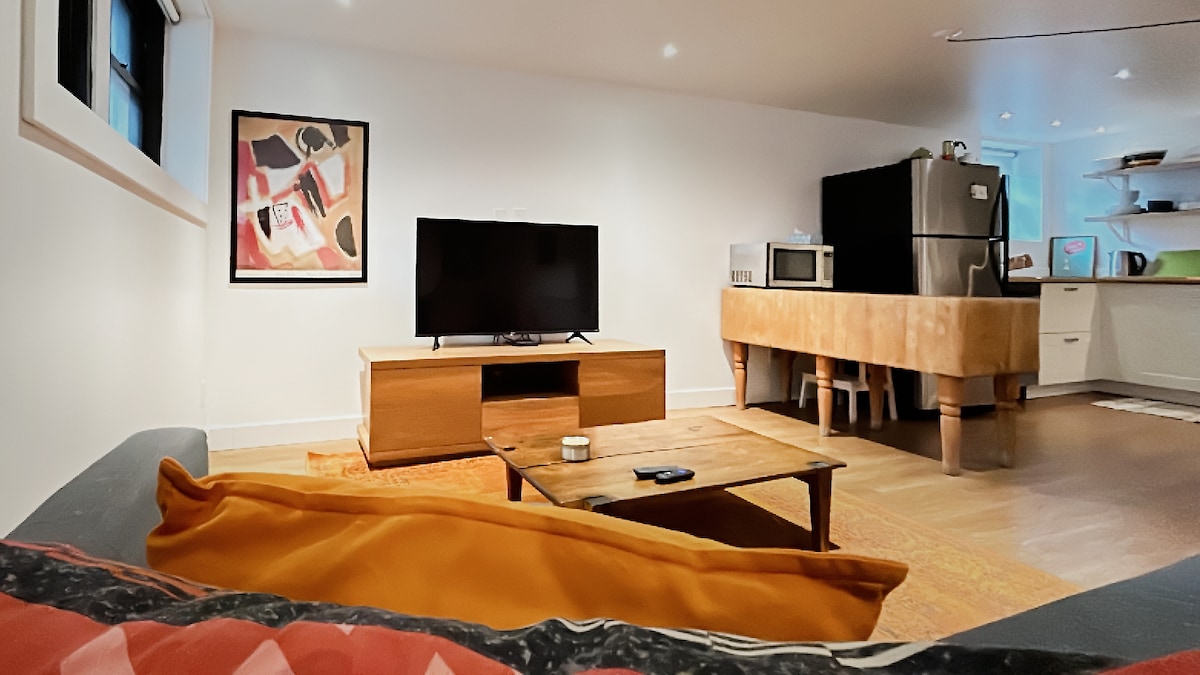 A comfortable living area features a flat-screen television resting on a wooden cabinet. A coffee table sits in front, accompanied by a cozy couch adorned with colorful cushions. The kitchen is visible in the background, revealing modern appliances and open shelving.