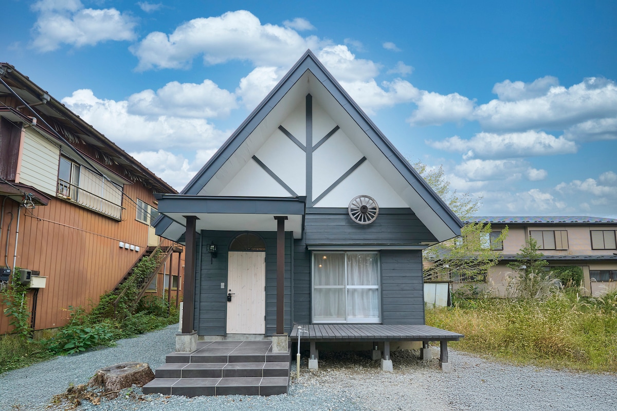 A charming house features a triangular roof and a large circular window. The entrance is accentuated by a welcoming porch, surrounded by a gravel path and greenery. Nearby structures provide context, while the cloudy blue sky adds to the ambiance.