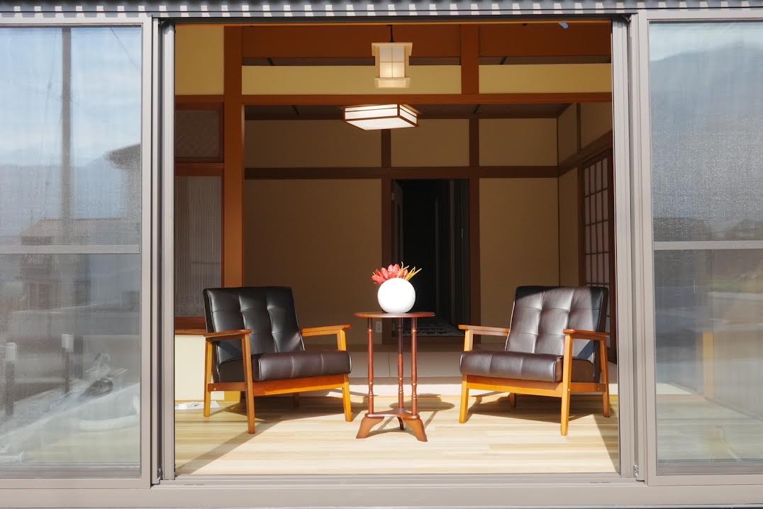 Two wooden armchairs are positioned facing each other on a light wooden floor, complemented by a small round table with a vase of fresh flowers. The sliding doors provide a seamless connection to the outdoor area, allowing natural light to fill the space.