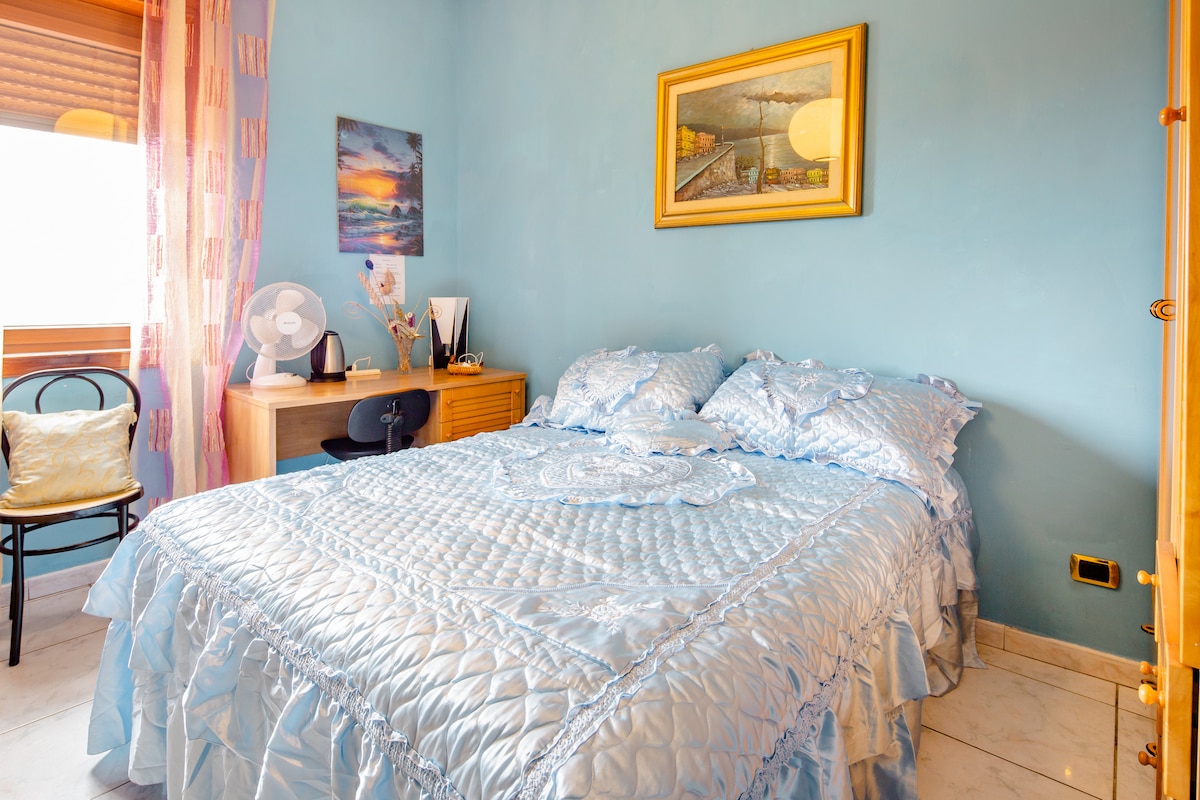 A spacious sleeping area features a blue-hued bedspread over a large bed, complemented by decorative pillows. A matching blue wall adds a soothing backdrop. A small wooden desk with a chair and a fan is positioned nearby, while artwork adorns the wall above the desk.