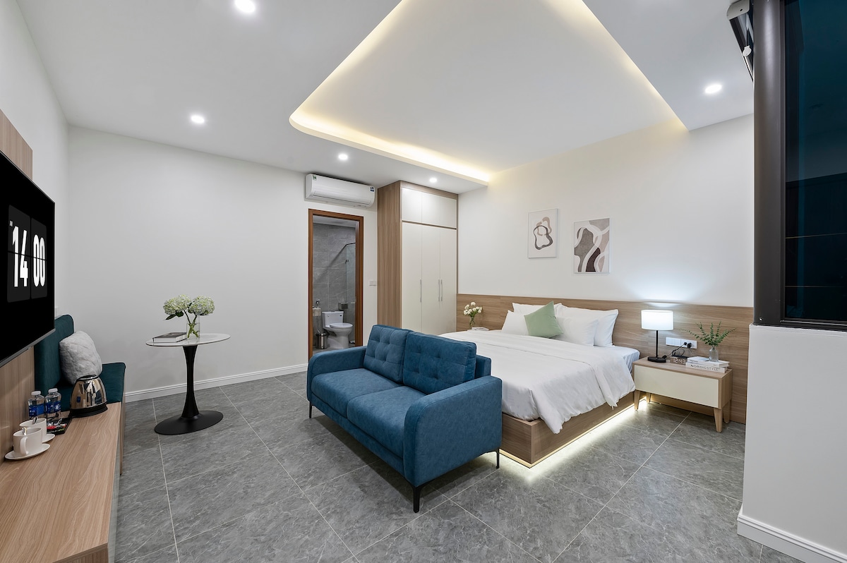 A modern room features a spacious bed with a soft white duvet and decorative pillows. A blue sofa is positioned nearby, accompanied by a small round table. Natural light fills the space, highlighting the sleek design and subtle decor elements.