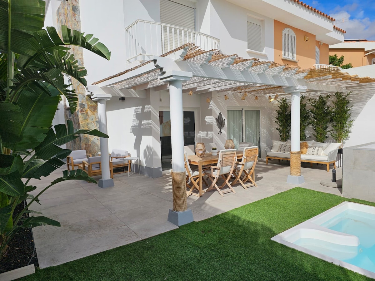 A covered outdoor dining area is depicted, featuring a wooden table surrounded by chairs. Lush green plants frame the space, while a private heated swimming pool is visible to the right. The patio is adorned with a blend of stone and white walls, creating a serene environment.