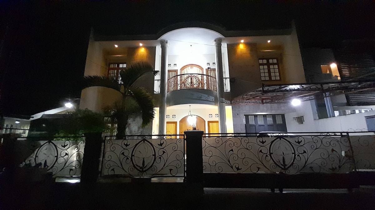 The exterior of a well-lit two-story home is showcased at night. A prominent balcony with decorative railings is visible above the entrance. Lush greenery and a patterned gate enhance the welcoming ambiance, while soft illumination highlights the architectural features.