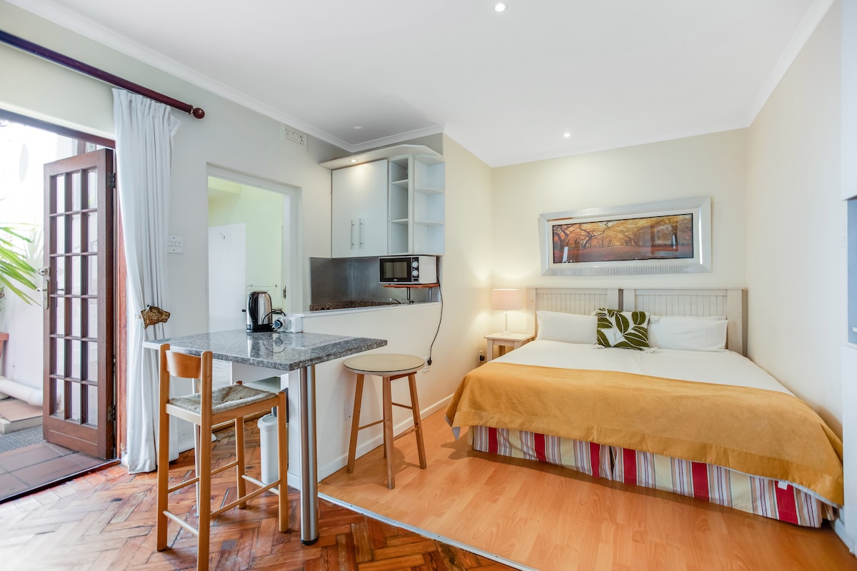 A compact living space features a spacious bed with a light beige blanket and striped bed skirt. A small dining area includes two wooden chairs and a granite-top table. Kitchen amenities are visible, and natural light fills the area from a nearby door opening to an outdoor space.