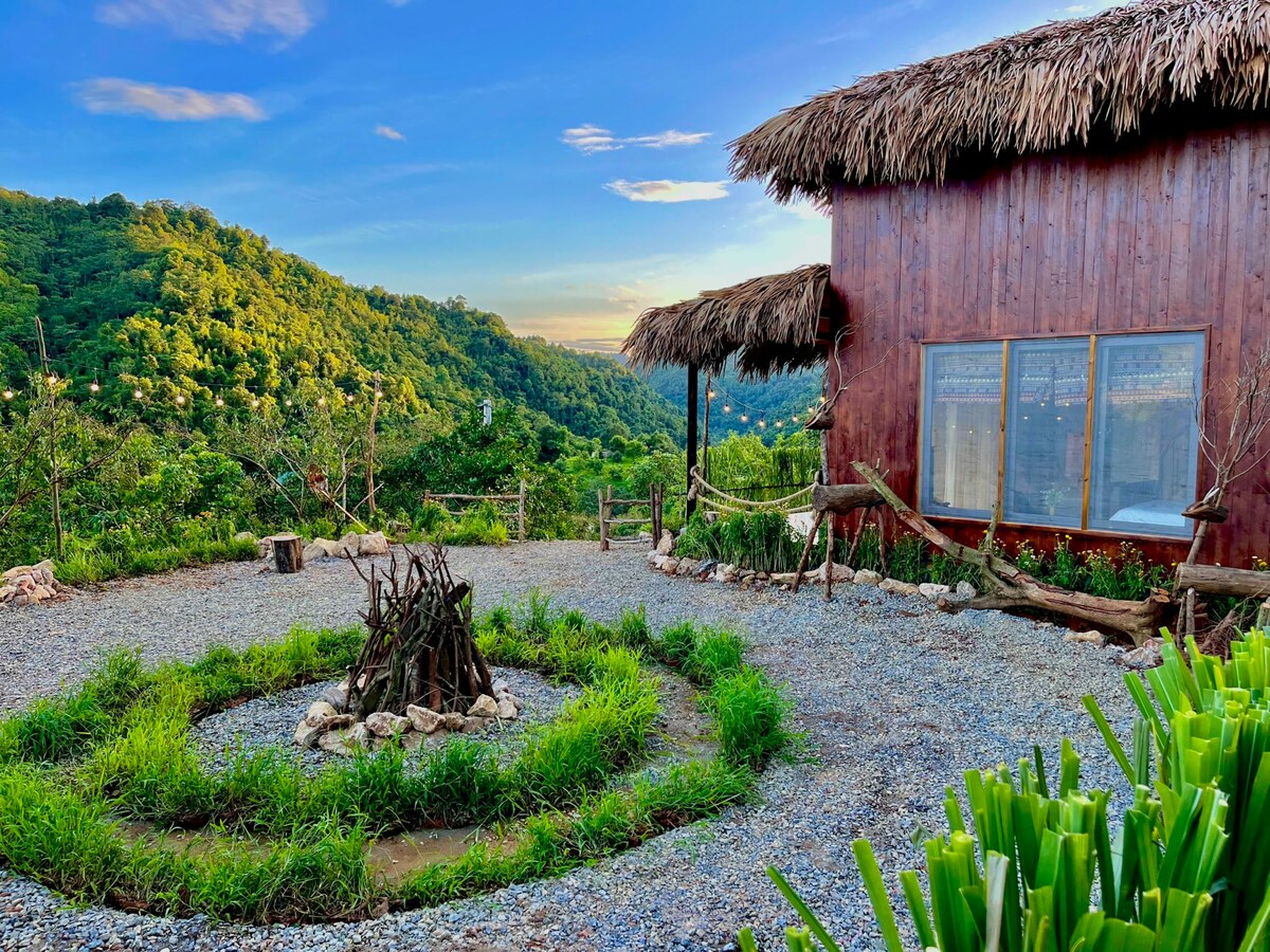 An outdoor area features a circular arrangement of stones and greenery surrounding a fire pit made of twigs. A wooden villa with a thatched roof is visible in the background, set against a hillside covered in lush trees and grasses.