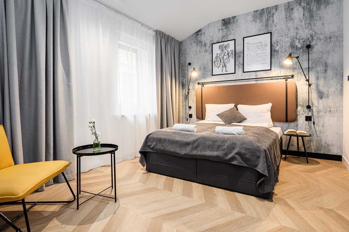 A modern room is seen featuring a spacious bed with a dark headboard, complemented by soft linens. Two decorative frames adorn the wall above the bed. A small round table and a yellow chair provide additional seating, illuminated by natural light through the sheer curtains.