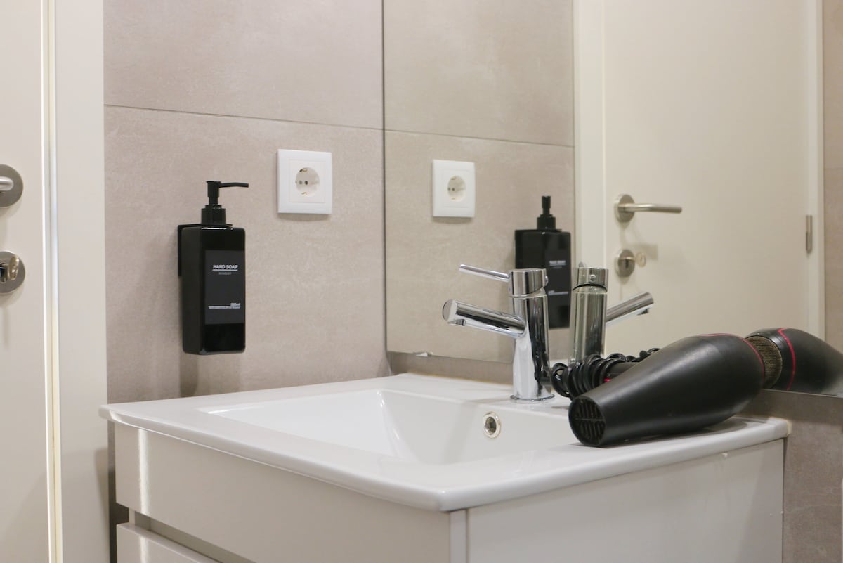 A modern bathroom sink is featured with a sleek, white countertop and a silver faucet. Wall-mounted dispensers are provided for amenities. A hairdryer rests beside the sink, and a clear mirror reflects the space, enhancing the overall clean and organized appearance.