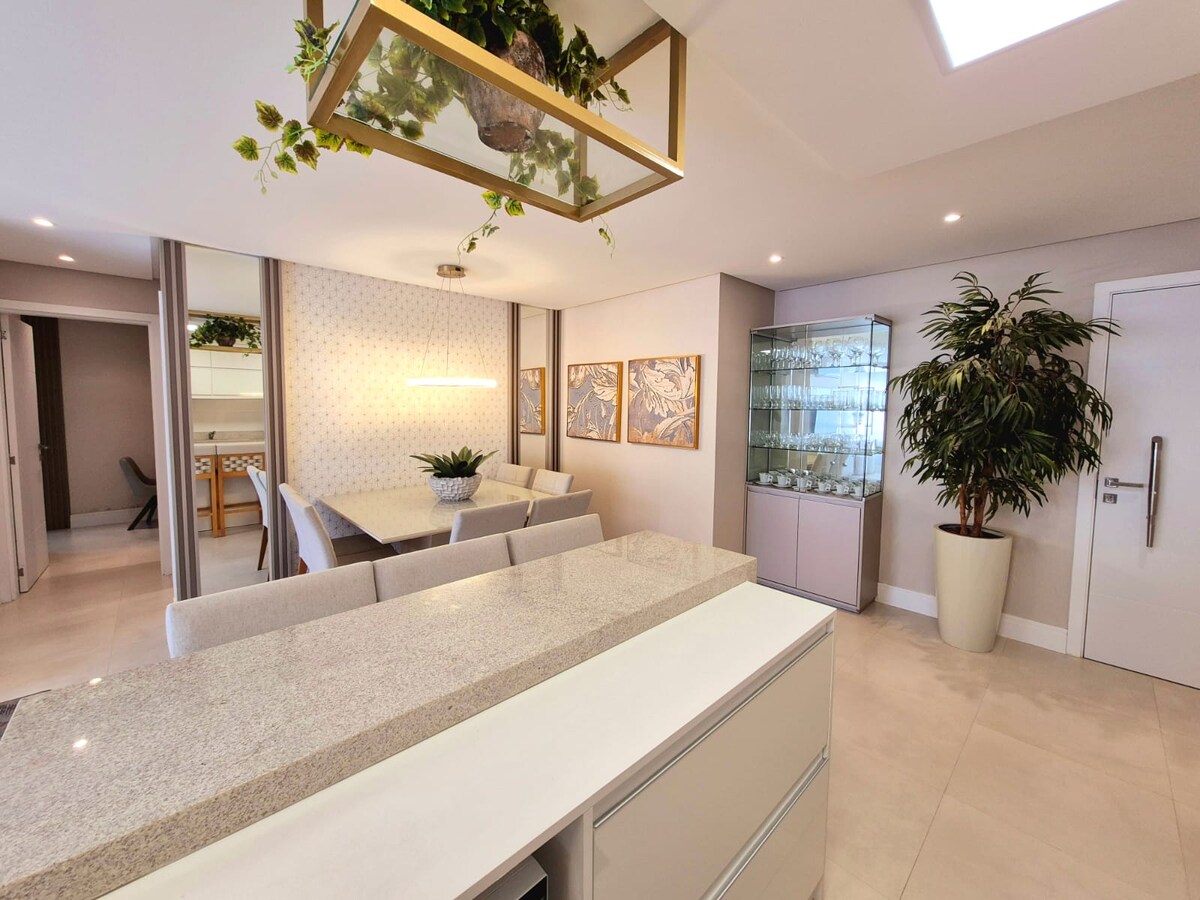 A modern living space features light-colored cabinetry and an elegant countertop. A dining area is visible, surrounded by tasteful decor and potted plants. Framed wall art adds a touch of sophistication, while a sleek glass display cabinet showcases assorted items.