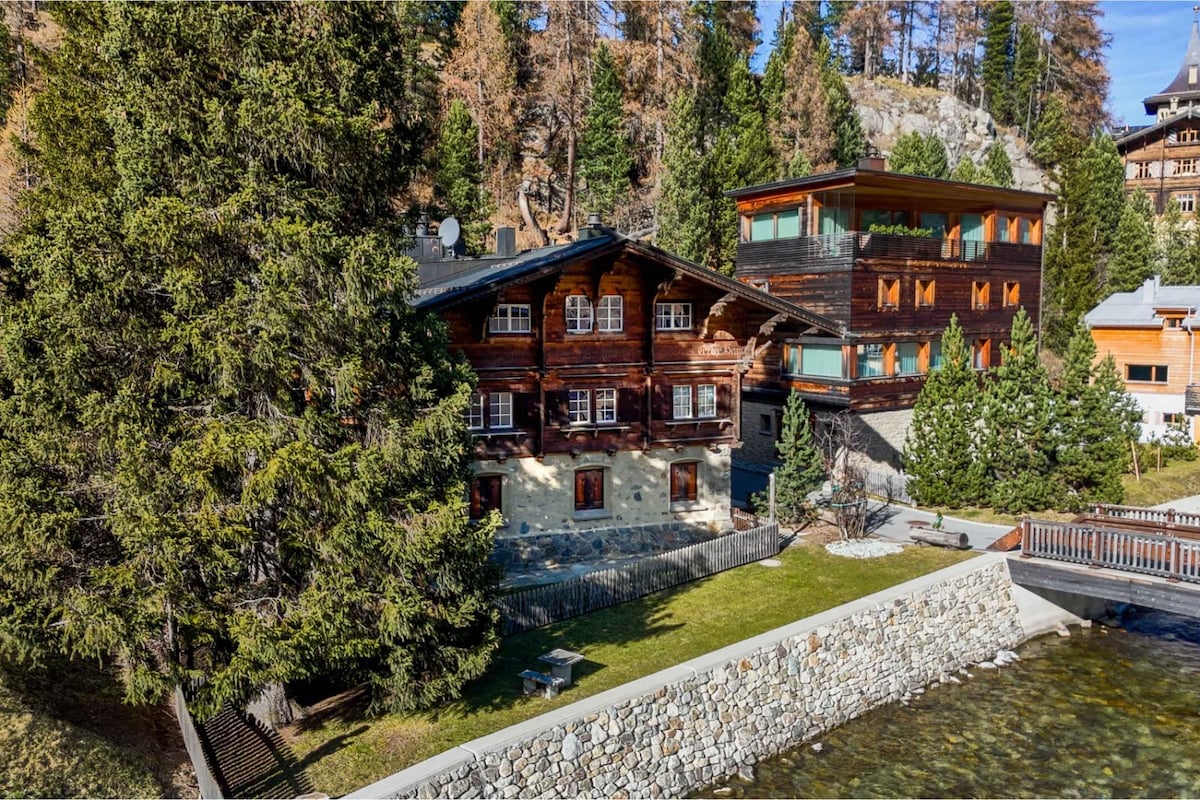 An alpine chalet with wooden exterior is nestled among tall trees, featuring a well-maintained lawn. A stone wall borders the property, while a picturesque stream runs nearby, enhancing the serene landscape. The building showcases multiple windows, allowing for ample natural light.
