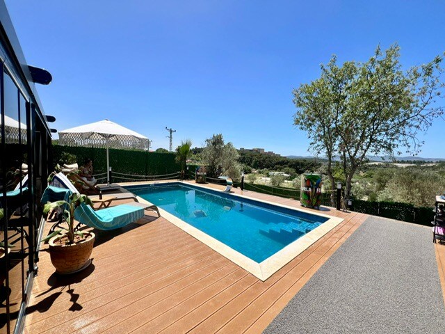 A spacious outdoor pool area is presented, featuring a rectangular pool surrounded by wooden decking. Comfortable loungers are positioned along one side, with parasols providing shade. Lush greenery and trees frame the scenic view, enhancing the serene atmosphere of the space.