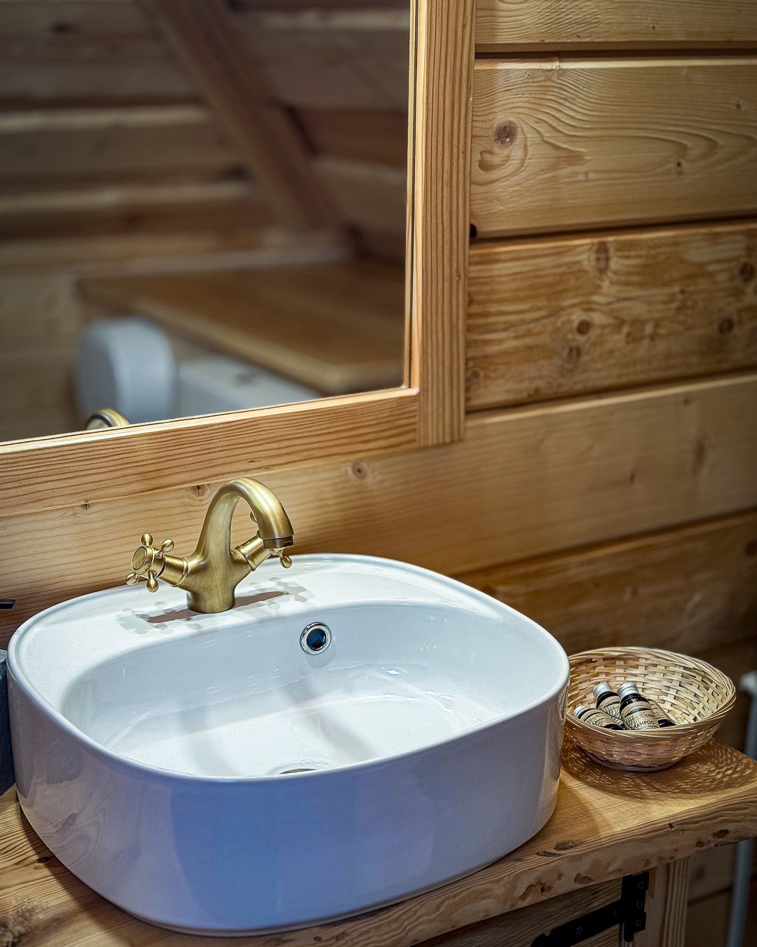 A modern sink is positioned on a wooden countertop, complemented by a stylish gold faucet. A mirror framed in natural wood hangs above, reflecting the cozy interior. A woven basket holds various toiletries, enhancing the space's rustic charm.