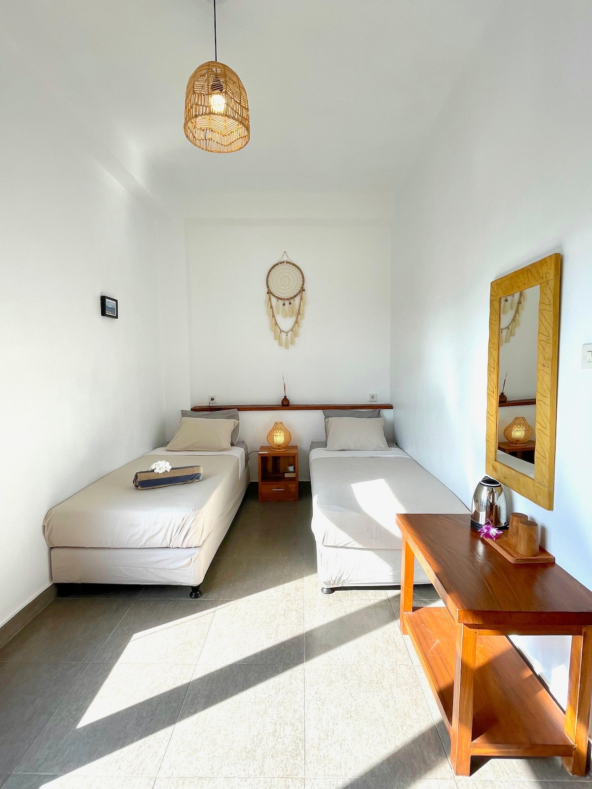 A bright room is shown featuring two single beds with light bedding positioned alongside a wooden nightstand. A woven pendant light hangs from the ceiling above, and a mirror is mounted on the wall opposite. A kettle is placed on a wooden table, inviting relaxation.