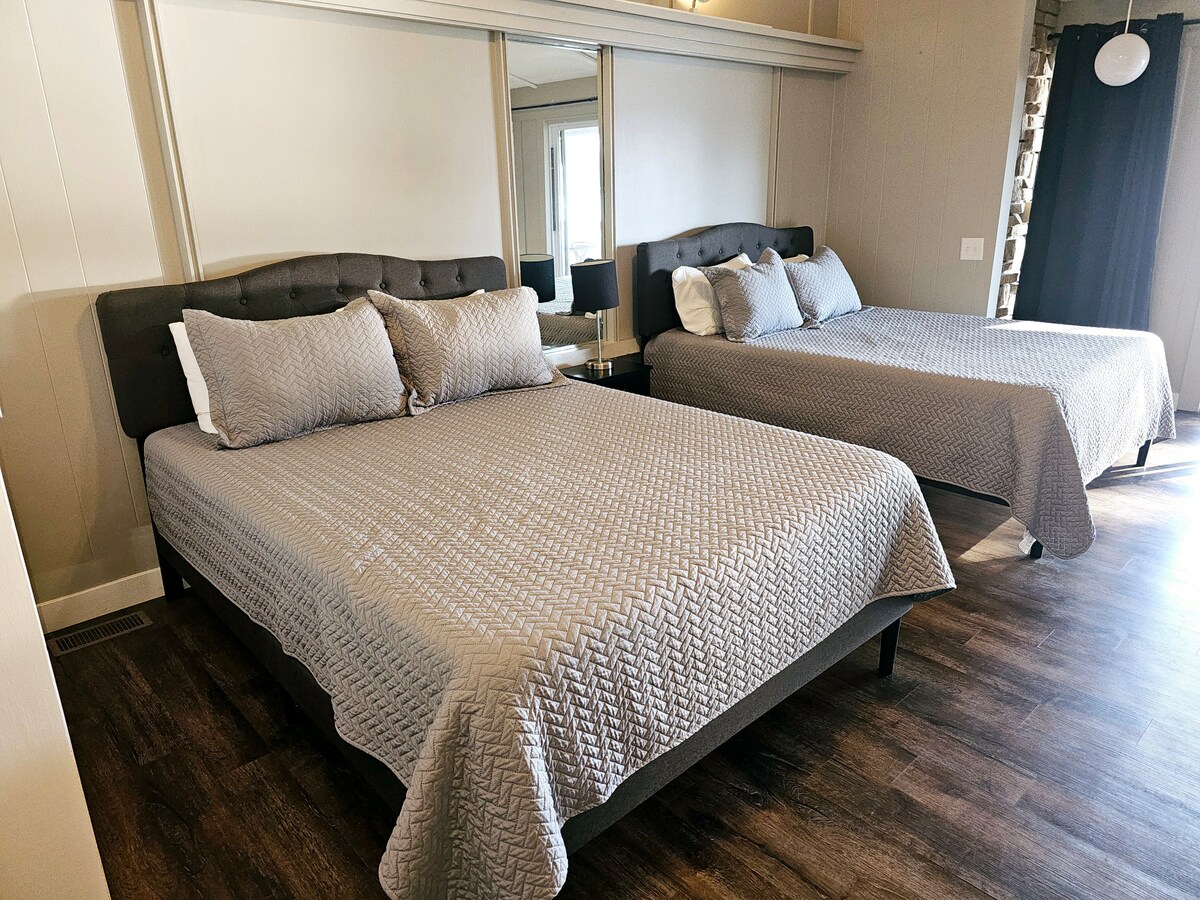 Two spacious beds are dressed in textured gray bedding, positioned within a well-lit room. A mirror spans the wall behind the beds, reflecting natural light, while earthy-toned flooring adds warmth to the space.
