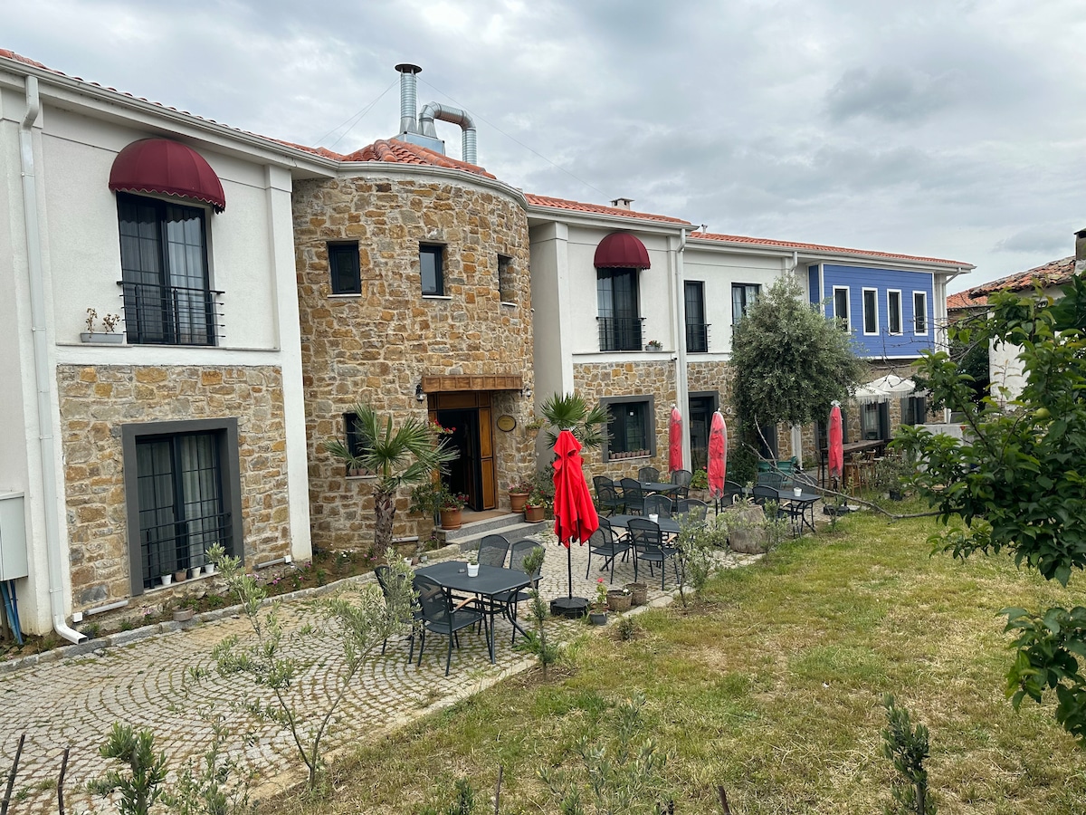 The exterior of a charming building is displayed, featuring a stone facade with large windows and red awnings. A spacious outdoor area is equipped with black metal tables and chairs, while red umbrellas provide shade. Lush greenery surrounds the setting, enhancing its inviting atmosphere.