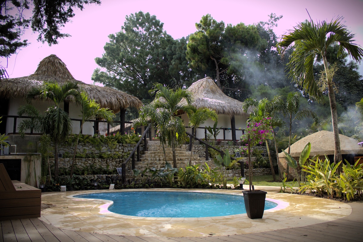 The outdoor area features a circular swimming pool surrounded by a wooden deck. Lush tropical plants and single-storied huts with thatched roofs create a natural setting. Stone steps lead to the structures, enhancing the scenic view. Soft smoke wafts from a nearby cooking area.