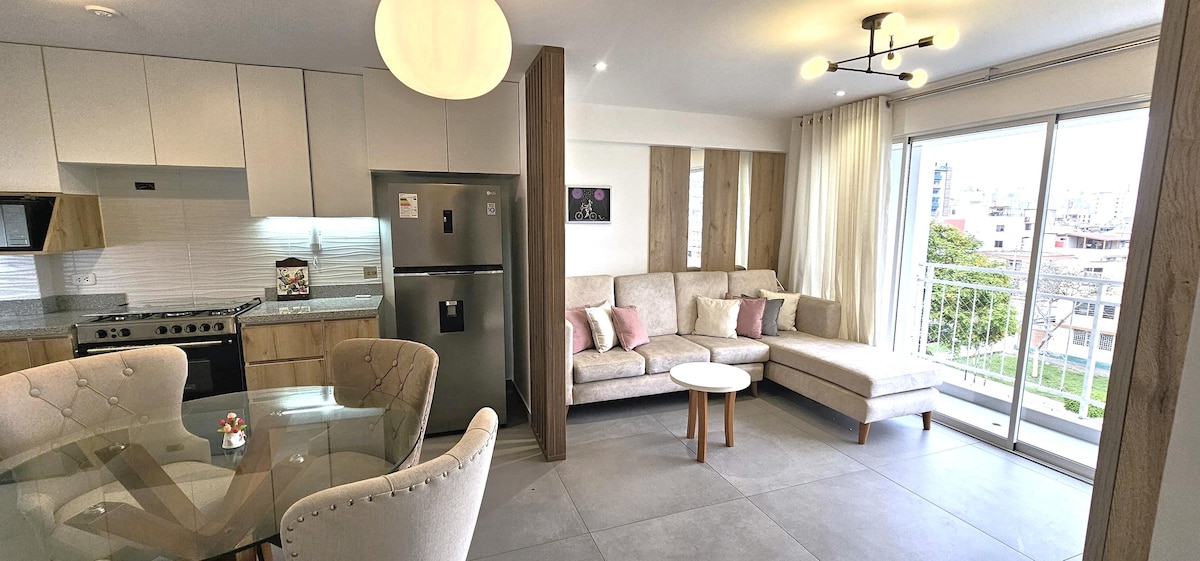 A modern living area features a comfortable sectional sofa with soft cushions, positioned near a balcony door that allows natural light to fill the space. A sleek dining table is visible, paired with elegantly upholstered chairs, creating a welcoming atmosphere in the open layout.