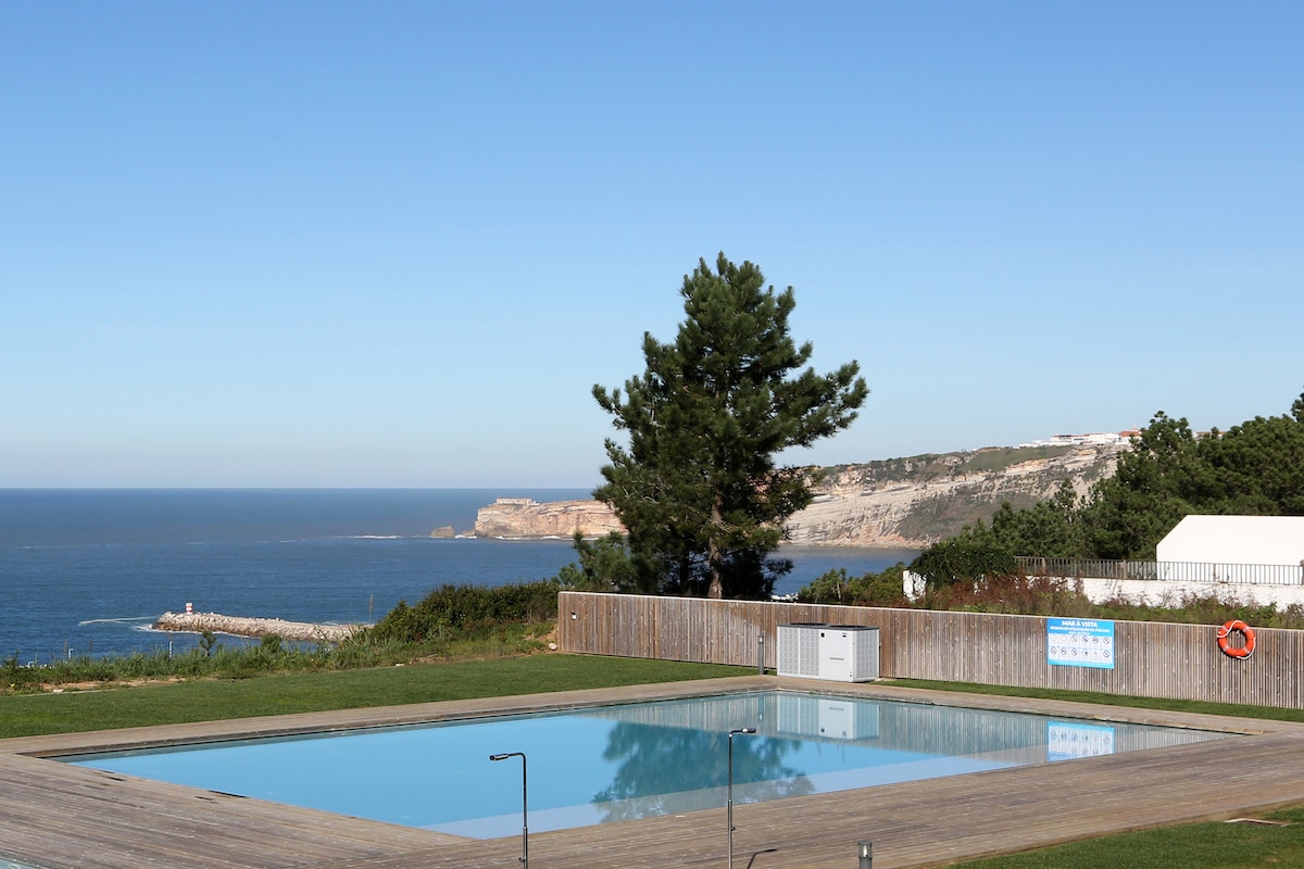 A tranquil outdoor swimming pool is set within a landscaped area, surrounded by wooden decking. The expansive view encompasses a serene coastline and lush greenery, with distant cliffs visible against a clear blue sky, creating a peaceful atmosphere.