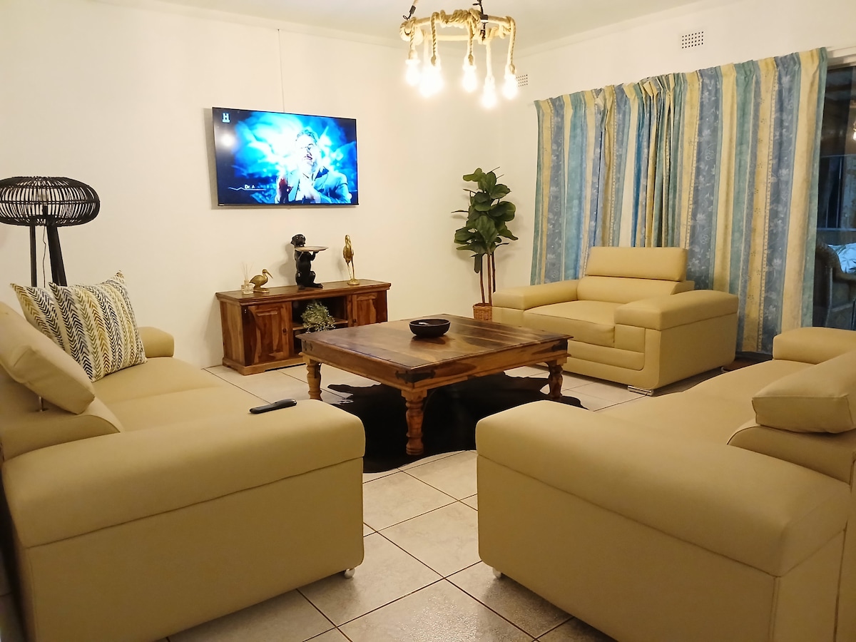 A welcoming living area features two light-hued sofas positioned around a wooden coffee table. A flat-screen television is mounted on the wall opposite, while patterned curtains frame the windows. A decorative plant and sculpture add a touch of elegance to the space.
