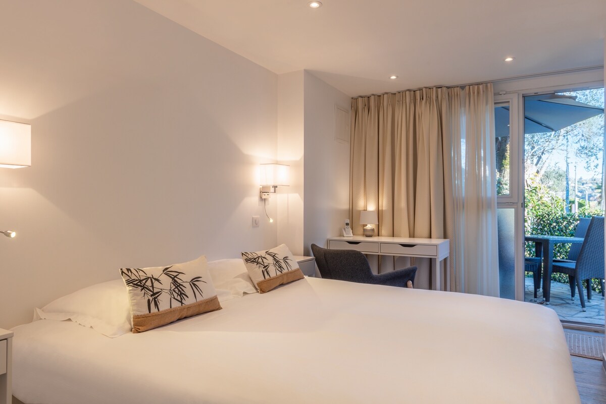 A spacious bedroom is furnished with a large bed featuring decorative pillows. Soft lighting enhances the inviting space. A desk and chair are positioned near sliding glass doors that lead to a private terrace, where outdoor seating can be seen.