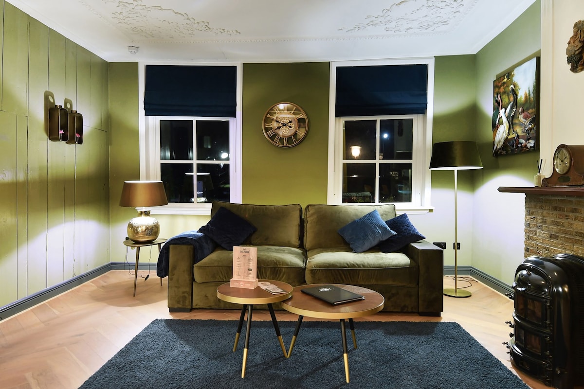 A cozy living area features a velvety sofa adorned with dark blue cushions, complemented by two round coffee tables. The green walls are accented by elegant wall decorations, and two large windows with dark blinds provide soft lighting, while a decorative fireplace adds character to the space.