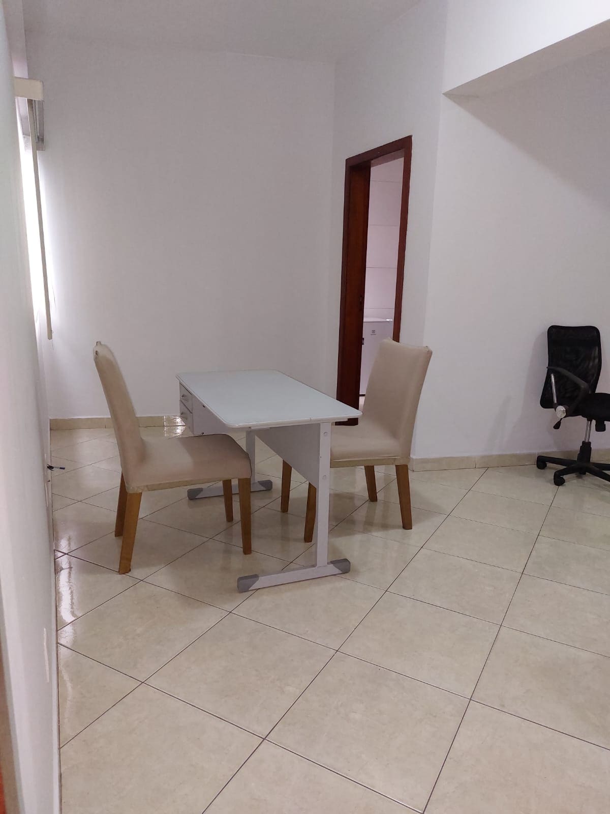 A small dining or workspace area is presented, featuring a rectangular white table paired with two light-colored chairs. A black ergonomic chair is visible nearby, on a tiled floor that reflects light. The walls are painted in a soft hue, contributing to a neat environment.