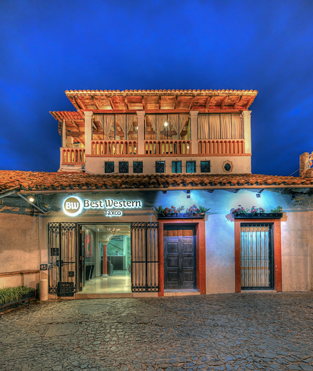 The exterior of Best Western Taxco is showcased under a deep blue sky, highlighting its charming architecture. The entrance is framed by traditional stone walls, with a wooden door adorned by decorative elements. Soft lighting accentuates the entrance, inviting guests to explore inside.