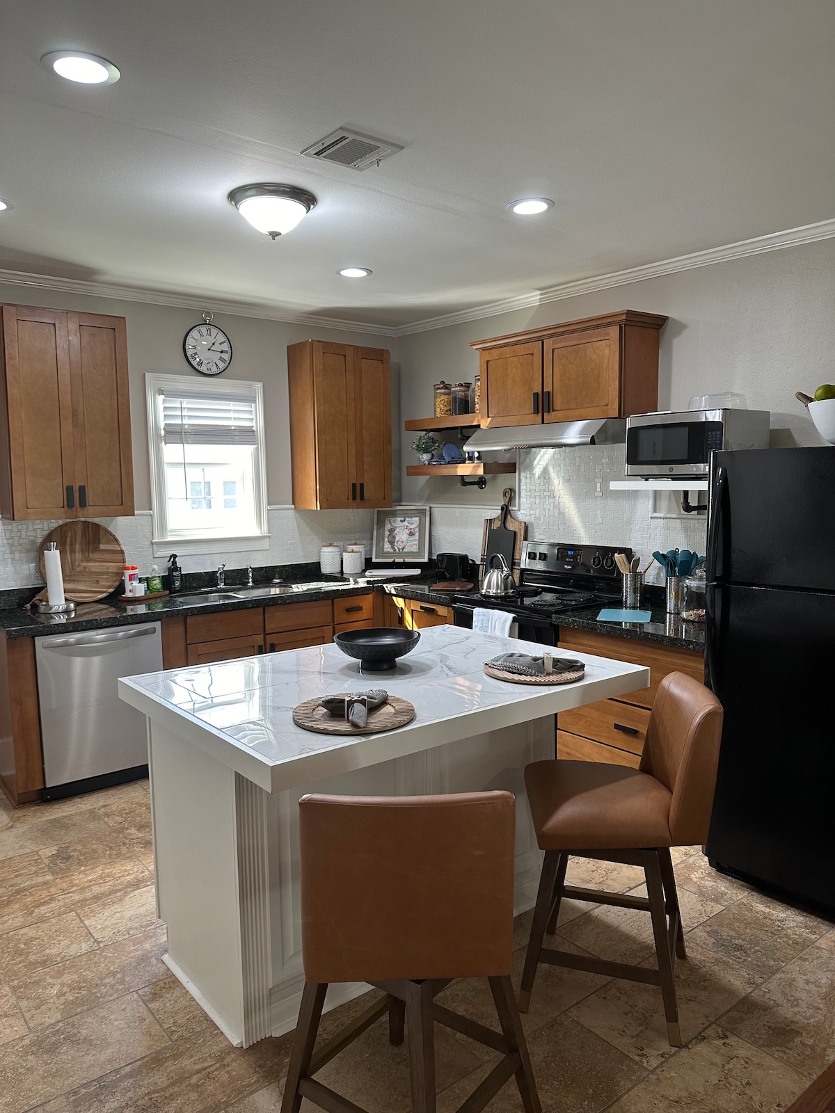 A modern kitchen features wooden cabinets, a sleek black refrigerator, and stainless-steel appliances. A central island with two brown chairs provides additional seating. Natural light enters through a window, illuminating the dark countertop and neatly arranged kitchen accessories.