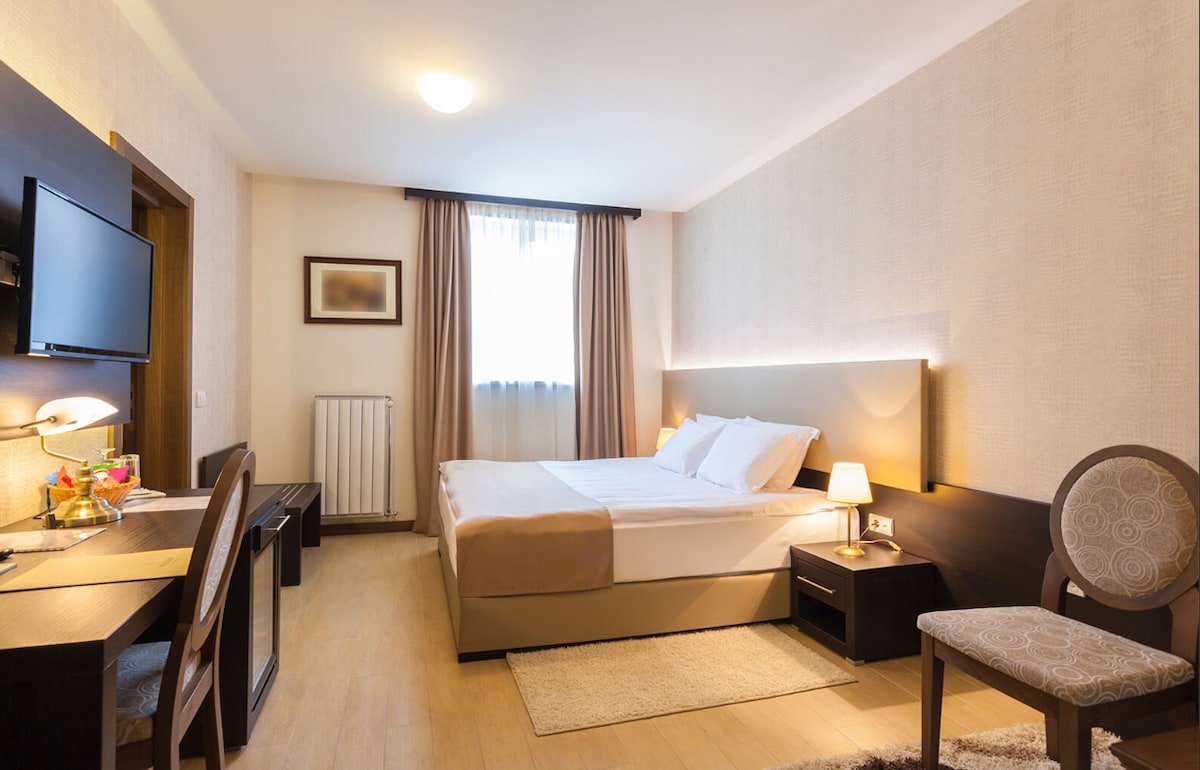 A comfortable guest room features a spacious bed with soft bedding and a modern headboard. A desk and chair are positioned against a wall, while a flat-screen television is mounted nearby. Natural light enters through a window dressed with light curtains.