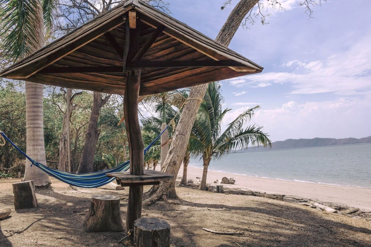 A shaded wooden structure provides a spot for relaxation, with a blue hammock hanging nearby. The sandy beach and calm water stretch into the distance, framed by palm trees and gentle waves lapping at the shore under a partly cloudy sky.