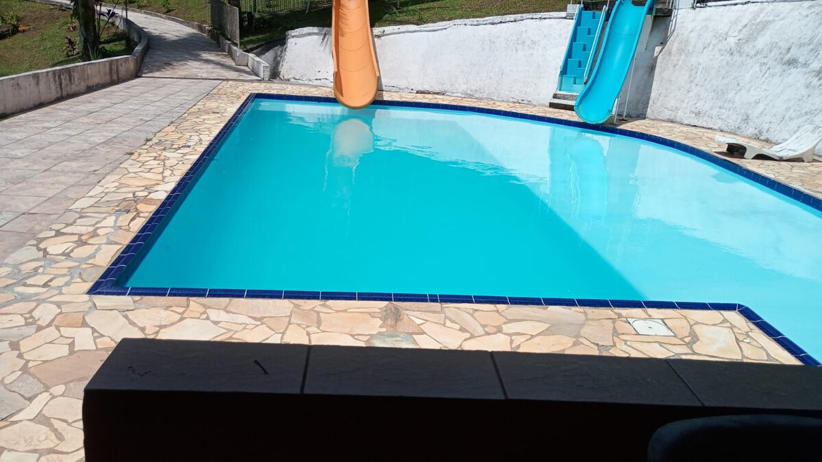 A clear swimming pool is surrounded by a textured stone patio. An orange water slide is positioned alongside the pool, which reflects the bright blue sky. Lush green grass is visible in the background, providing a natural contrast to the pool area.