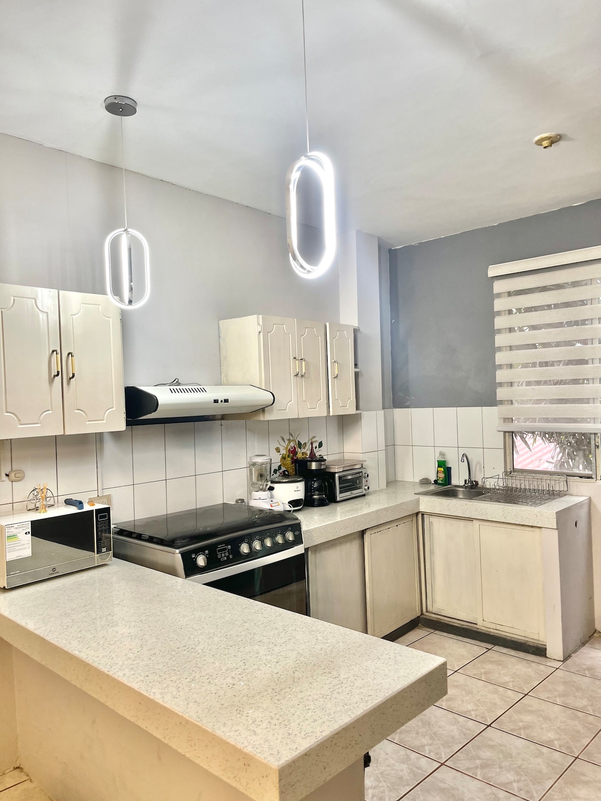 A modern kitchen is presented with white cabinetry and a light-colored countertop. The space is equipped with a stovetop, oven, and microwave. Pendant lighting is featured above the counter, and a window with shades illuminates the area.